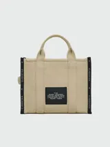 Bolso tipo tote de lona color negro con letras blancas bordadas en el frente que dicen 'THE TOTE BAG MARC JACOBS'. Cuenta con asas superiores cortas y una correa de hombro ajustable y extraíble.
