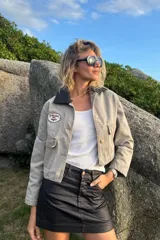 Campera corta de jean color visón, con cuello desmontable de tela engomada, cierre frontal y bolsillos plaqué con solapa en el pecho. Cuenta con un parche bordado en el delantero.