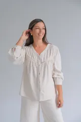 Blusa de lino color beige, con cuello en V, cierre frontal con botones y detalle de alforzas y encaje en el pecho. Presenta mangas largas con puños sencillos y un corte holgado.
