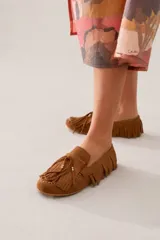 Mocasín de gamuza color tostado con diseño de flecos en todo el contorno y en la capellada. Presenta un cordón decorativo con detalles metálicos dorados en el frente.