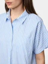 Camisa de popelina con mangas cortas y cuello clásico. Presenta cierre frontal de botones y diseño estampado de rayas verticales blancas y celestes.