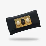 Cartera tipo sobre de yute negro con solapa, placa metálica dorada grabada con tres piedras ovaladas, dos blancas en los extremos y una oscura en el centro. Incluye correa de cadena desmontable.