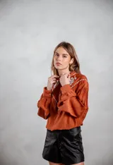 Camisa de tela texturizada color terracota, con mangas largas abullonadas y detalle de bordado blanco en el canesú delantero. Presenta cierre frontal con botones.