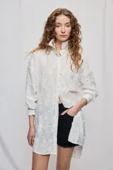 Vestido camisero corto y de silueta amplia en color blanco, con cuello clásico, cierre frontal de botones y mangas largas. Cuenta con diseño bordado floral en toda la prenda y dobladillo asimétrico, más largo en la parte trasera.
