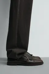 Zapato de vestir estilo monk strap fabricado en piel color marrón oscuro. Presenta ajuste mediante dos hebillas laterales metálicas, detalle de flecos en el empeine y puntera con perforaciones decorativas tipo brogue.