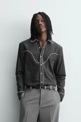 Camisa de corte relajado en color negro con rayas verticales finas. Presenta un diseño estilo western con vivos blancos en contraste en el canesú, cuello, tapeta frontal y puños. Cuenta con cuello solapa, cierre frontal de botones y mangas largas.