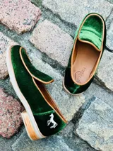Mocasines de terciopelo verde oscuro, con ribetes en tono tostado y detalle de aplique metálico de un caballito en el talón. Poseen suela y taco bajo de madera.