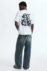 Remera de algodón blanca de corte holgado, con cuello redondo y un pequeño estampado gráfico negro en el pecho con el texto "NEXT LEVEL READY".