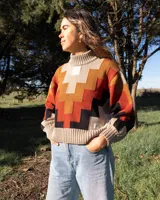 Buzo de tejido de punto con cuello alto y diseño geométrico en zigzag. Presenta una combinación de colores cálidos como naranja, rojo, negro y beige, con puños y cintura acanalados.
