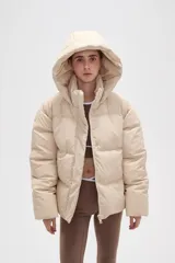 Campera puffer color beige con diseño capitoneado, capucha integrada y cierre frontal oculto. Presenta bolsillos laterales y un corte holgado.