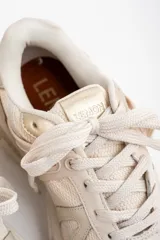 Zapatilla deportiva de diseño urbano en tonos neutros, combinando paneles de malla, gamuza y detalles metalizados en color dorado. Cuenta con suela robusta y acordonado frontal.