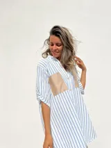 Camisola de corte oversize con estampado de rayas verticales azules sobre fondo blanco. Presenta cuello camisero, cierre frontal con botones y dos bolsillos frontales en contraste de color beige con acabado tipo gamuza.