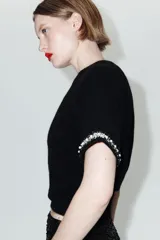 Jersey de punto negro de cuello redondo y manga corta, con detalle de perlas decorativas en el borde de las mangas.