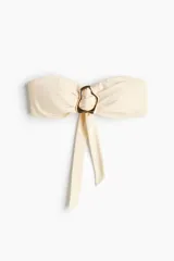Top de bikini strapless de color beige, con copas acolchadas, varillas laterales y ribete de silicona en el escote. Cuenta con un detalle metálico dorado en el centro con tiras colgantes y ajuste en la espalda.