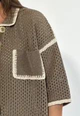 Chaqueta de punto calado en color negro con detalles de bordes en color crudo. Presenta cuello camisero, cierre frontal con botones, mangas cortas y un bolsillo frontal tipo parche.