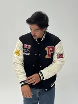 Campera estilo varsity con cuerpo negro y mangas blancas. Presenta parches bordados en el pecho y las mangas, cierre frontal con botones a presión y puños, cuello y ruedo acanalados con rayas blancas y negras.