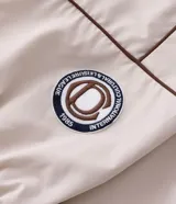 Campera de tejido técnico color beige con detalles de costuras en contraste color marrón. Presenta cuello alto, cierre frontal con cremallera, mangas largas con puños elásticos y estampa frontal con texto. Incluye un detalle bordado en la manga.