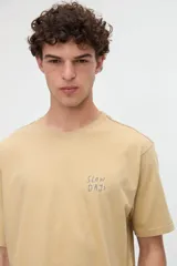 Remera de manga corta color beige, confeccionada en jersey de algodón con textura suave y calce relaxed. Presenta costuras a contratono en la espalda y un diseño estampado en la parte posterior con ilustraciones de estilo lineal y frases manuscritas.