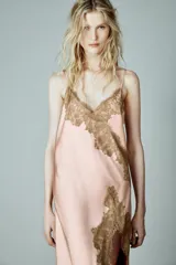 Vestido largo tipo lencero de satén color rosado, con escote en pico y tirantes finos. Presenta aplicaciones de encaje a contraste en el escote y en el lateral, con un diseño de bajo asimétrico.