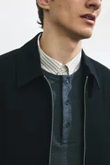 Campera negra de tejido suave, corte estándar, con cuello clásico, cierre frontal y bolsillos laterales con cierre. Presenta puños abotonados y elástico oculto en la parte posterior del bajo.