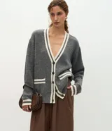 Cardigan de punto gris con escote en V, cierre frontal con botones y detalles de vivos en color crema en el escote, puños, bolsillos y ruedo. Presenta un diseño de corte holgado con mangas largas y bolsillos funcionales.