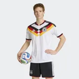 Camiseta de fútbol Adidas de la primera equipación de Alemania, modelo 26. Diseño principal blanco con un patrón en V en el pecho que incorpora los colores de la bandera alemana (negro, rojo y amarillo/dorado). Posee cuello en V con ribete rojo y detalles en las mangas. El escudo de la federación alemana con cuatro estrellas está bordado en el pecho, junto al logo de Adidas.