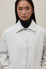 Chaqueta larga color beige, con cuello camisero y cierre frontal de botones a presión. Tiene dos bolsillos laterales y está confeccionada con poliamida y poliéster 100% reciclados.