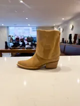 Bota de caña alta estilo western confeccionada en cuero vacuno color suela. Presenta un diseño versátil con estribo desmontable y flecos decorativos en la zona del tobillo, taco de madera y punta fina.
