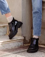 Botinetas de caña baja con suela track gruesa, combinando cuero negro en la puntera, gamuza marrón en el lateral y detalles de textura animal print tipo serpiente en tonos marrones. Presenta un cierre de solapa con velcro en el empeine.