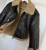 Campera tipo gamulán de cuero sintético negro con interior y cuello de corderito sintético en color beige. Presenta cierre frontal, bolsillos laterales con cierre, cierres en los puños y detalles de hebillas metálicas en el cuello.
