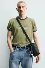 Bolso tipo bandolera de diseño minimalista en color negro, con cierre de cremallera superior y asa de hombro ajustable. Incluye un detalle decorativo de charms metálicos en uno de los laterales.