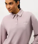 Buzo deportivo de corte oversized color lila, con cuello polo y cierre frontal con botones a presión. Presenta mangas largas con puños ajustados y una estampa bordada en el pecho. Confeccionado en una mezcla de poliéster, modal y elastano.