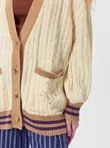 Cardigan de punto trenzado en color negro con detalles en color crema en los bolsillos, puños y ruedo. Presenta un diseño oversize, cierre frontal con botones y bordado decorativo en uno de los bolsillos delanteros.