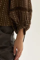 Blusa de color marrón con estampado de rayas verticales y pequeñas flores blancas. Presenta cuello redondo con lazo de ajuste, cierre frontal con botones y mangas largas abullonadas con detalles de encaje calado y puños ajustables con lazo.