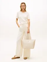 Cartera tipo tote color beige con acabado liso, doble asa superior de cinta y logotipo metálico dorado de la marca en la parte frontal. Cuenta con compartimiento principal con cierre de cremallera.