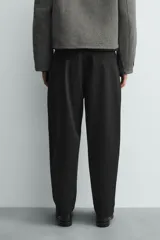 Pantalón negro de corte relaxed fit y estilo wide leg, con pliegues frontales dobles, bolsillos delanteros y detalle de bolsillos traseros de vivo. Cierre frontal cruzado con cremallera y gancho metálico oculto.