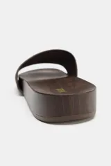 Sandalia plana con tira ancha de gamuza marrón y suela con efecto madera. Presenta detalle de tachas metálicas en el lateral y punta redonda.