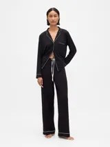 Pijama de dos piezas confeccionado en modal negro, compuesto por una camisa de manga larga con cuello solapa, cierre frontal de botones y vivos en contraste blanco, y un pantalón de corte recto con cintura elástica ajustable mediante cordón y vivos en el ruedo.