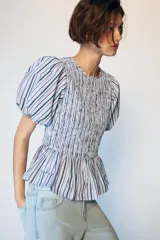 Top de cuello redondo con cuerpo de nido de abeja elástico y mangas globo con elástico en los puños. Presenta un diseño a rayas verticales en tonos azul, blanco y granate, con un bajo estilo peplum.