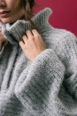 Buzo tejido en mohair de color gris, con silueta amplia y corte en línea A. Presenta cuello tipo remera con broches que permiten cerrarlo como polera, mangas largas con puños y un diseño de tejido horizontal.