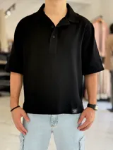 Chomba oversize negra de lino, con cuello polo, cierre frontal con botones y mangas cortas. Presenta un pequeño parche con logo en el borde inferior.