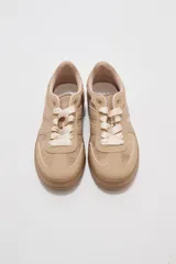 Zapatillas urbanas de corte bajo en tonos beige y tostado, combinando paneles de cuerina y gamuza sintética. Cuentan con cordones anchos tipo cinta de raso en color crudo, pespuntes decorativos y suela de goma al tono.