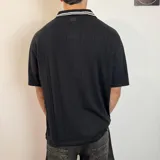 Chomba de tejido de punto color negro, con cuello polo y cierre frontal parcial. Presenta diseño de rayas verticales sutiles en el tejido, detalles de líneas blancas en los hombros y estampado frontal con texto y gráficos en color blanco.