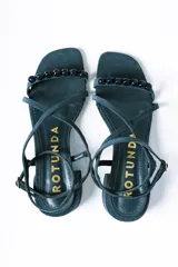 Sandalias de taco cuadrado medio, confeccionadas en gamuza color azul. Presentan tiras finas que cruzan el empeine, una pulsera ajustable al tobillo y detalle de cuentas decorativas en la tira frontal.