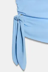 Top celeste de popelina sin mangas, con cuello barco y botones dorados en el hombro. Presenta diseño fruncido en los laterales y detalle de nudo con tiras en la cintura.