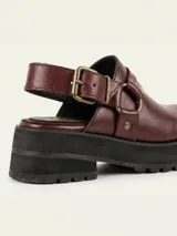 Zueco de cuero vacuno color bordó, con diseño de punta redondeada, detalle de tira con tachas metálicas y herrajes en tono oro viejo. Cuenta con un taco cuadrado de 4,5 cm y suela de cuero.