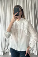 Camisa blanca de textura arrugada con cuello alto, volados frontales y detalle de plisado en el pecho. Presenta cierre frontal con botones y mangas largas con puños terminados en volados.