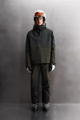Cazadora deportiva de esquí con diseño bicolor en tonos negro y verde oliva. Presenta capucha amplia con visera, cierre frontal con cremallera termosellada, bolsillos frontales y en manga con cierres impermeables, y aberturas de ventilación en las axilas. Incluye faldón interior antinieve, puños ajustables y tecnología de rescate RECCO.