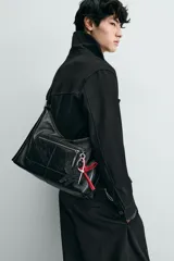 Bolso tipo bandolera de cuero sintético negro con acabado brillante. Presenta un compartimento principal con cierre de cremallera, un bolsillo frontal con cierre y detalle de charms colgantes decorativos. Incluye asa de hombro ajustable.
