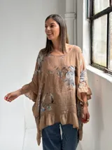 Blusa holgada color camel con estampado floral en tonos grises y azules, cuello redondo y mangas amplias con volados, al igual que el ruedo inferior.